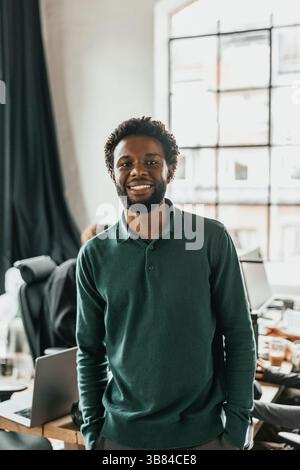 Portrait d'un homme aux cheveux bouclés souriant professionnel de L'INFORMATIQUE debout avec les mains dans les poches dans le bureau Banque D'Images