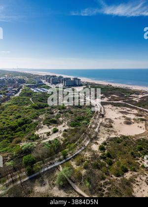 Vue aérienne d'une zone côtière avec des dunes de sable, une végétation verdoyante, des sentiers sinueux et des bâtiments modernes en bord de mer, le tout s'étendant le long du rivage Banque D'Images