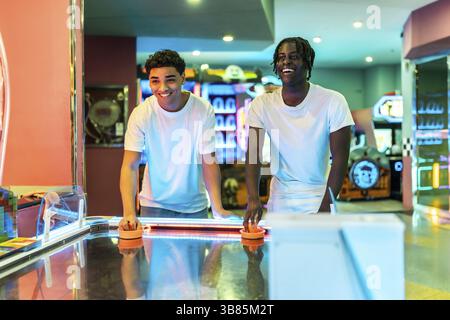 Deux jeunes hommes profitant d'un week-end ensemble, jouant au hockey pneumatique dans une salle de jeux très éclairée Banque D'Images