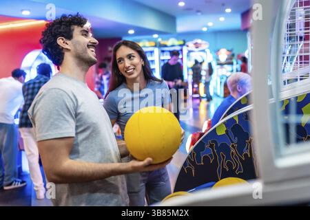 Jeune couple multiethnique s'amusant à jouer au basket-ball d'arcade ensemble, profitant de leur temps de loisirs de week-end dans un centre de divertissement Banque D'Images
