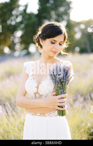 Sibenik, Croatie - 05.06.17: Mariée dans une robe de dentelle blanche a ses bras croisés sur sa poitrine et tient un bouquet de lavande en eux, regardant Banque D'Images