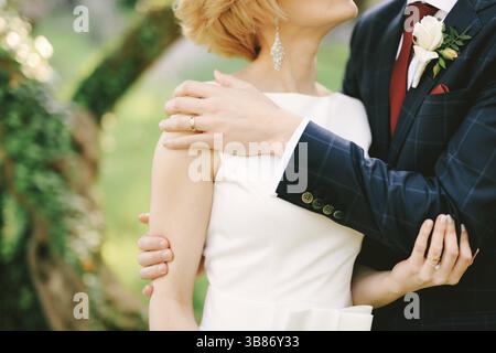 Groom dans un costume à carreaux bleu embrasse la mariée dans une robe blanche dans le parc sur le fond d'un arbre vert entrelacé avec du lierre. Gros plan. Banque D'Images