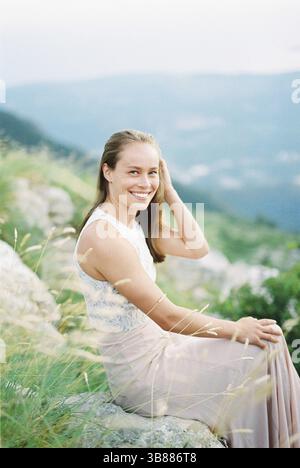 Fille souriante redresse ses cheveux avec sa main tout en étant assise sur une pierre dans les montagnes. Banque D'Images