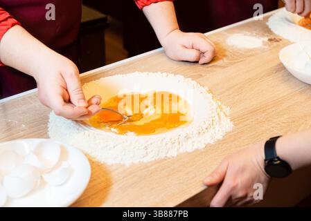 Les mains commencent à mélanger les œufs crus dans un puits de farine sur une table en bois, en commençant le processus de fabrication des pâtes dans un cours de cuisine. Banque D'Images