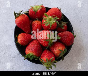 Vue de dessus d'un bol noir rempli de fraises fraîches mûres, placé sur une surface légère à la texture rugueuse. Concept de nourriture fraîche et saine. Banque D'Images