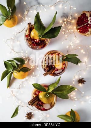 Sangria d'hiver sur fond de marbre blanc. Sangria de vacances en verres avec tranche de fruits, grenade et épices sur décoration chaîne d'éclairage de table. Banque D'Images