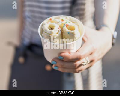 Crème glacée roulée dans la tasse de cône dans les mains de la femme. Femme en robe rayée tient une tasse de cône avec la crème glacée roulée de banane kiwi de style thaïlandais Banque D'Images
