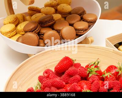 Un assortiment de macarons colorés dans un bol, accompagnés de fraises fraîches et de framboises, parfait pour un affichage de dessert vibrant lors d'une célébration Banque D'Images