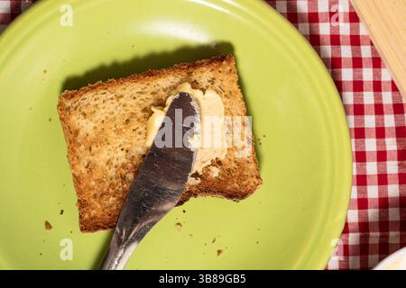 Couteau à beurre étalant de la margarine sur du pain grillé pour le petit déjeuner sur une assiette verte et une nappe à carreaux rouge Banque D'Images