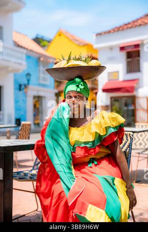 Afro-Latina Palenquera femme en costume coloré rouge, vert et jaune assis avec bol de fruits sur la tête à Carthagène, Colombie. Palenqueras Banque D'Images