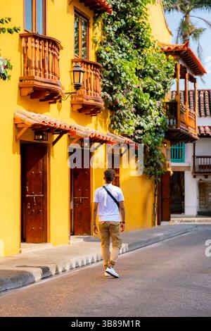 Jeune homme latino méconnaissable en t-shirt blanc et pantalon kaki se promène détendu le long d'une rue jaune vibrante à Carthagène, en Colombie. Banque D'Images