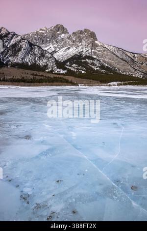Un lac gelé calme s'étend au premier plan, présentant des configurations de glace complexes sous un ciel crépusculaire. Des montagnes majestueuses s'élèvent en arrière-plan, Banque D'Images