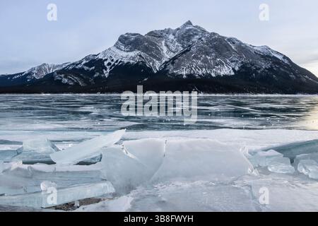 Des morceaux de glace gelés sont dispersés le long de la rive d'un lac, encadrés par une chaîne de montagnes majestueuse sous un ciel nuageux. L'atmosphère hivernale souligne Banque D'Images