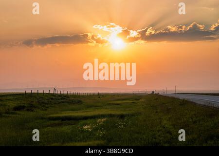 La lumière dorée se propage à travers l'horizon alors que le soleil se couche le long d'une autoroute tranquille. Les champs verdoyants encadrent la scène, améliorant l'atmosphère paisible de Banque D'Images