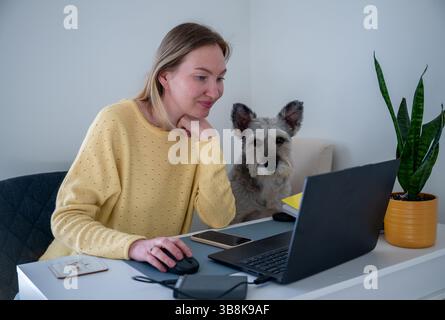 Femme en pull jaune travaillant sur ordinateur portable à la maison avec son chien, concept de travail à distance Banque D'Images