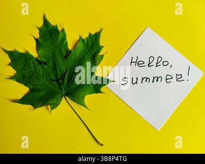 Feuille d'érable verte unique et feuille de papier blanche avec l'inscription HELLO, SUMMER. Composition sur fond jaune. Copier l'espace. Gros plan. Banque D'Images