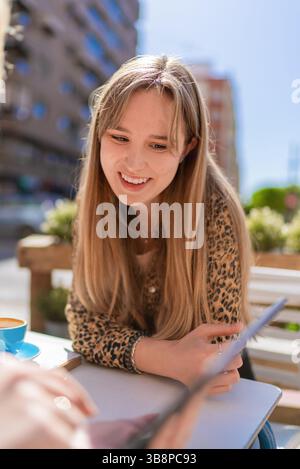 Femme blonde en chemise imprimé léopard se relaxant à la terrasse du bar extérieur, sirotant un café et souriant tout en parcourant le menu Banque D'Images