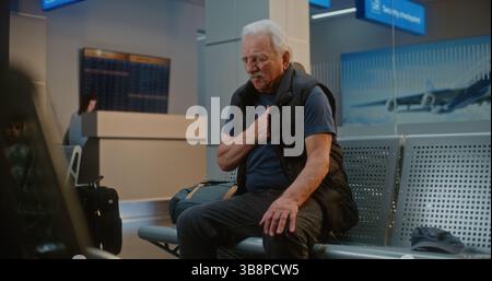 Terminal de l'aéroport international bondé : homme senior ayant une crise cardiaque, serrant la poitrine dans la douleur assis dans le salon des départs du hub aérien. Personnes diverses et personnel de l'aéroport aidant les passagers âgés. Banque D'Images