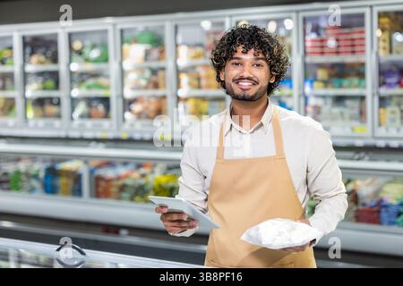 Un employé souriant d'une épicerie tient un paquet de nourriture tout en regardant la caméra. Il se tient près d'un congélateur. Banque D'Images
