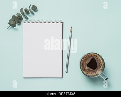 Carnet en papier vierge avec crayon et tasse à café. Carnet de croquis en papier vide avec pochoir et branches d'eucalyptus sur fond bleu. Vue de dessus ou plan de travail. Banque D'Images