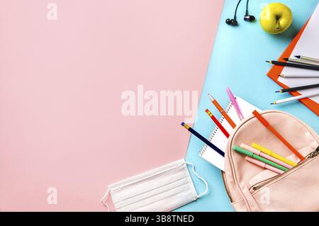 Bureau étudiant créatif avec masque de protection, fournitures scolaires modernes colorées et sac à dos rose sur fond bleu et rose duotone.Vue de dessus.Pose à plat. Banque D'Images