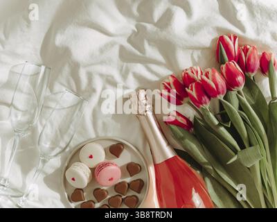 Bouquet de tulipes rouges, vin rose, verres à champagne, macarons et bonbons en forme de coeur sur draps blancs. Concept de la Saint-Valentin, date, mars 8. Ressort Banque D'Images
