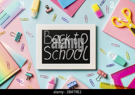 Tableau noir avec lettres Retour à l'école fait avec de la craie.Fournitures scolaires, crayons, ciseaux et livres de couleur sur fond bleu et rose.Vue de dessus ou Banque D'Images