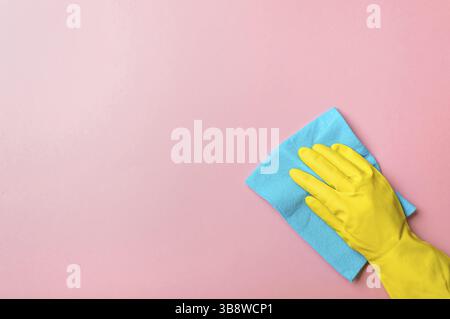 Main de l'employé dans un gant de protection en caoutchouc jaune tenir un chiffon bleu essuyant un fond de mur rose. Service de nettoyage de maison, concept de nettoyage général ou régulier. Banque D'Images