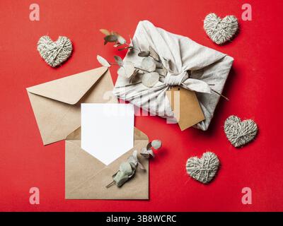 Zero Waste concept de Saint-Valentin et maquette sur rouge. Emballage en tissu cadeau écologique dans le style Furoshiki, enveloppe en papier artisanal, carte de voeux vide Banque D'Images