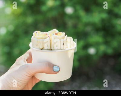 Glace roulée dans une tasse conique dans la main de femme. Femme tient une coupe conique avec de la glace de style thaïlandais kiwi banana roulé. Extérieur. Lumière naturelle espace de copie pour Banque D'Images