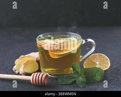 Tisane au gingembre, à la menthe, au citron et au miel. Tasse en verre à thé chaud avec vapeur et ingrédients sur fond de ciment noir. Copier l'espace Banque D'Images