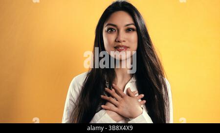 Jeune femme asiatique exprimant sa gratitude avec les mains sur la poitrine sur fond jaune Banque D'Images