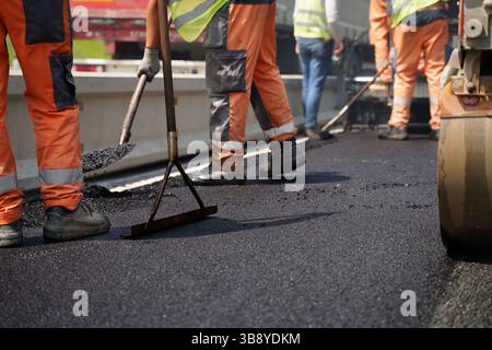 Prise de vue à faible angle de plusieurs jambes de travailleurs de la construction utilisant des outils pour étaler de l’asphalte frais à côté d’une barrière routière, mettant en évidence le travail d’équipe et la précision Banque D'Images