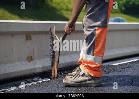 Prise de vue à faible angle des jambes d’un travailleur de la construction à l’aide d’un outil pour étaler de l’asphalte frais à côté d’une barrière routière, mettant en évidence la précision de la construction routière Banque D'Images