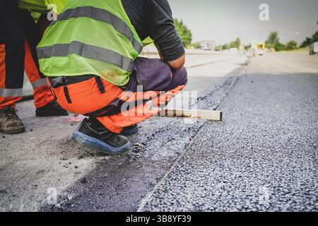 Travailleur de la construction mesurant l’asphalte frais pour s’assurer de son niveau, assurant précision et qualité pendant le processus de pavage routier sur un chantier de construction Banque D'Images