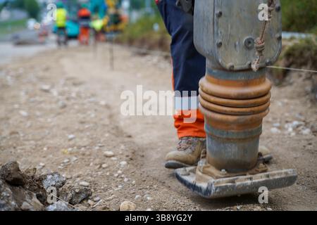 Gros plan des jambes des travailleurs à l'aide d'une plaque de construction vibrante pour comprimer et niveler le gravier, en préparant une surface lisse pour la nouvelle couche d'asphalte sur un Banque D'Images