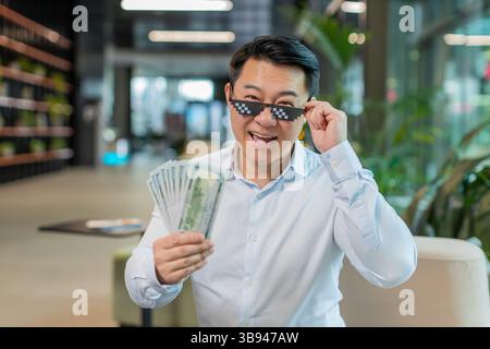 Heureux riche homme d'affaires chinois mûr portant des lunettes de soleil Thug Life montrant des billets de banque d'argent dollar assis dans le hall moderne du bureau. Carrière d'affaires de succès, gagnant de loterie, grand revenu, richesse. Banque D'Images