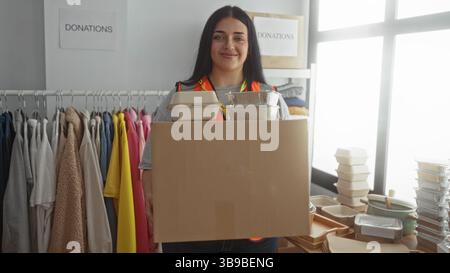 Une femme bénévole porte une boîte dans un centre de dons caritatifs entouré de vêtements et de contenants alimentaires à l'intérieur. Banque D'Images