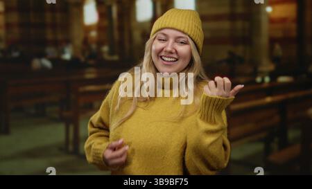 Femme à l'église souriant joyeusement, faisant des gestes de guitare, portant un pull jaune et un bonnet dans un cadre intérieur avec des bancs, créant un chaud et chee Banque D'Images