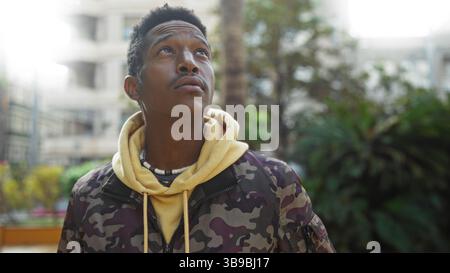 Jeune homme dans le parc urbain regarde avec une expression réfléchie, entouré de verdure et de paysage urbain, portant un sweat à capuche décontracté et veste camouflage suggérant un Banque D'Images