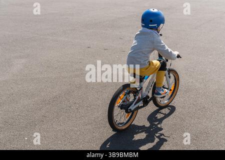 Un enfant portant un casque fait du vélo sur un espace extérieur pavé. L'image capture un moment de mise au point et d'exploration. Entouré de verdure et ouvert Banque D'Images