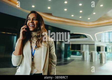 Un avocat indien confiant se tient dans un lobby d'entreprise moderne, s'engageant dans un appel téléphonique avec le sourire. Elle respire le professionnalisme et un comportement positif Banque D'Images