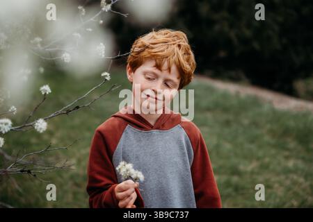 Garçon roux souriant dans le jardin de printemps tout en tenant des fleurs blanches Banque D'Images