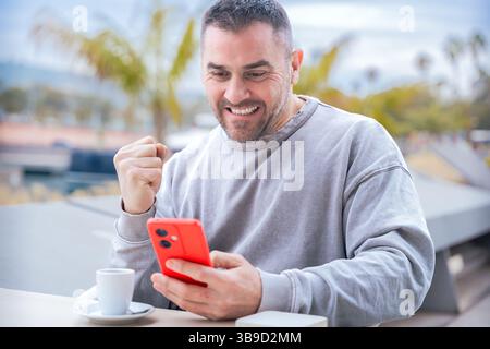 Homme célébrant après avoir gagné le pari de jeu en ligne en utilisant smartphone au café en plein air Banque D'Images