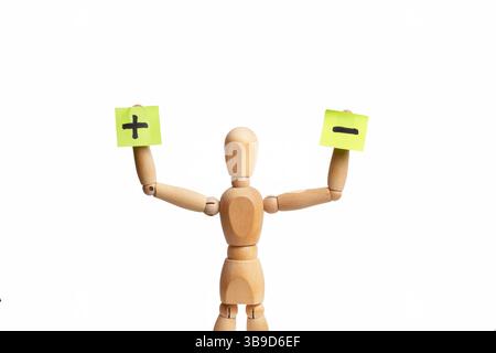 Un jouet mannequin en bois tenant du papier collant avec des signes moins et plus dans sa main sur fond blanc Banque D'Images