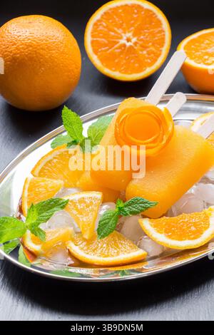 Crème glacée sorbet orange décorée d'une fleur d'écorce d'orange, feuilles de menthe sur plaque d'argent avec glaçons, tranches d'orange près des oranges sur fond noir. Crème glacée sorbet à l'orange popsicles. Vertical. Banque D'Images