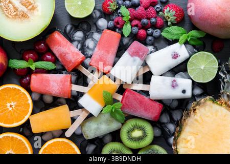 Les pop de crème glacée colorés faits avec diverses baies sont placés à plat sur une surface élégante. Autour d'eux se trouvent des fruits frais comme le kiwi, les oranges et les melons, améliorant l'ambiance estivale. Banque D'Images