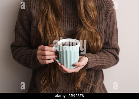 Femme tenant une tasse pastel. Matin d'automne confortable à l'intérieur. Boisson chaude à la main. Pull marron et cheveux longs. Moment paisible de style de vie hygge. Banque D'Images