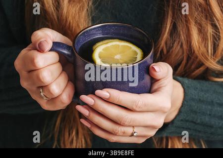 Thé chaud avec tranche de citron. Mains tenant le mug bleu. Moment de boisson confortable. Boisson chaude hivernale. Femme appréciant le thé. Détente et confort. Banque D'Images