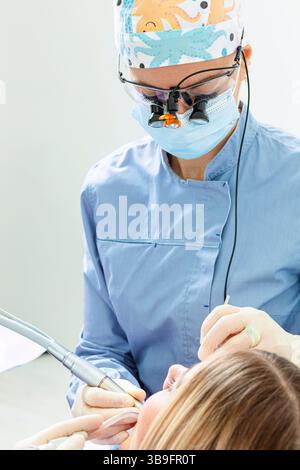 Dentiste portant des loupes et un masque chirurgical opérant sur les dents du patient à l'aide d'une perceuse dentaire et d'un éjecteur de salive Banque D'Images